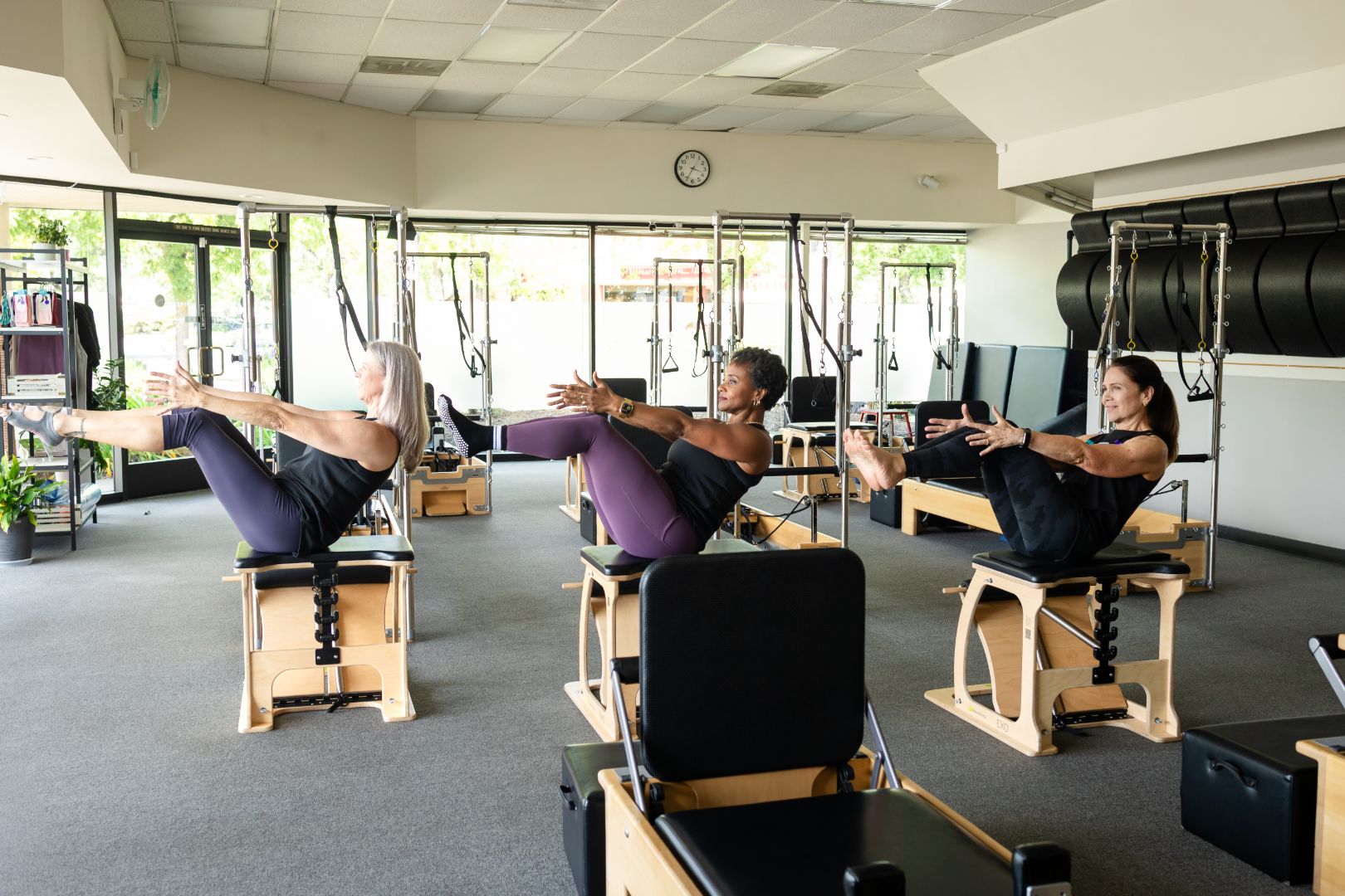 Three women perform Pilates teaser exercises on reformer chairs.